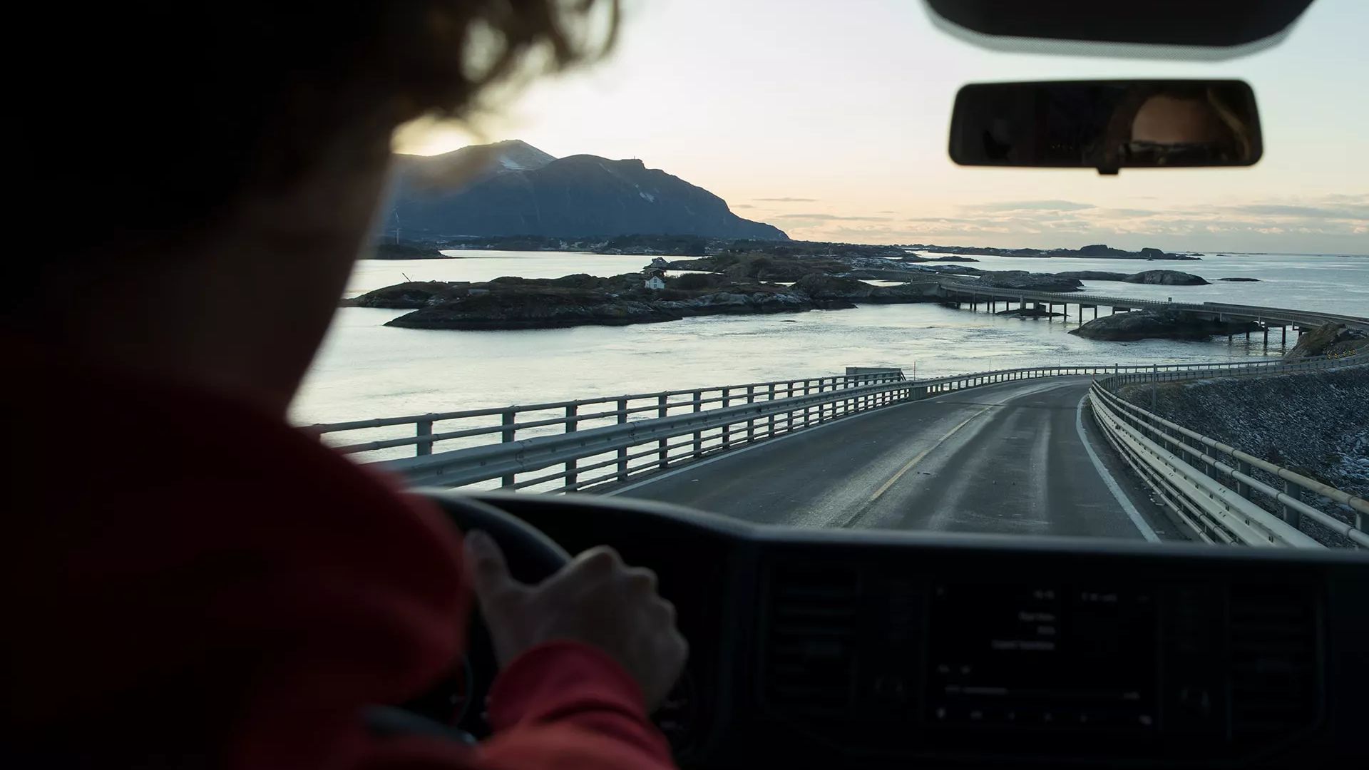 Eine Person fährt auf einer Küstenstraße bei Sonnenuntergang. Die Straße schlängelt sich vorbei an Wasser und Bergen. Der Blick aus dem VW Grand California zeigt teils bedeckte Hintergründe und ein spiegelndes Meer.