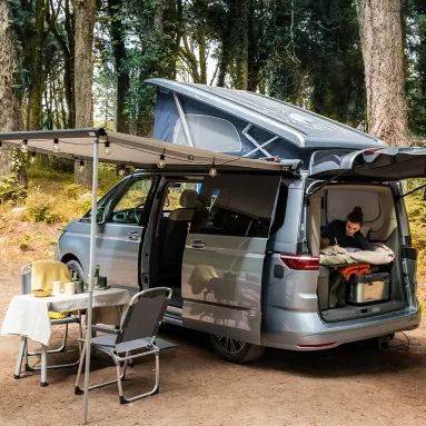 Ein Campingbus steht im Wald, das Dach ist aufgestellt und die Seitentür sowie die Heckklappe sind offen. Vor dem Bus sind ein Tisch und ein Stuhl aufgebaut. Es deutet auf ein gemütliches Campingerlebnis im Grünen hin.