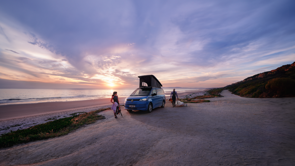 Ein blauer Volkswagen California mit aufgestelltem Dach steht an einem Strand. 