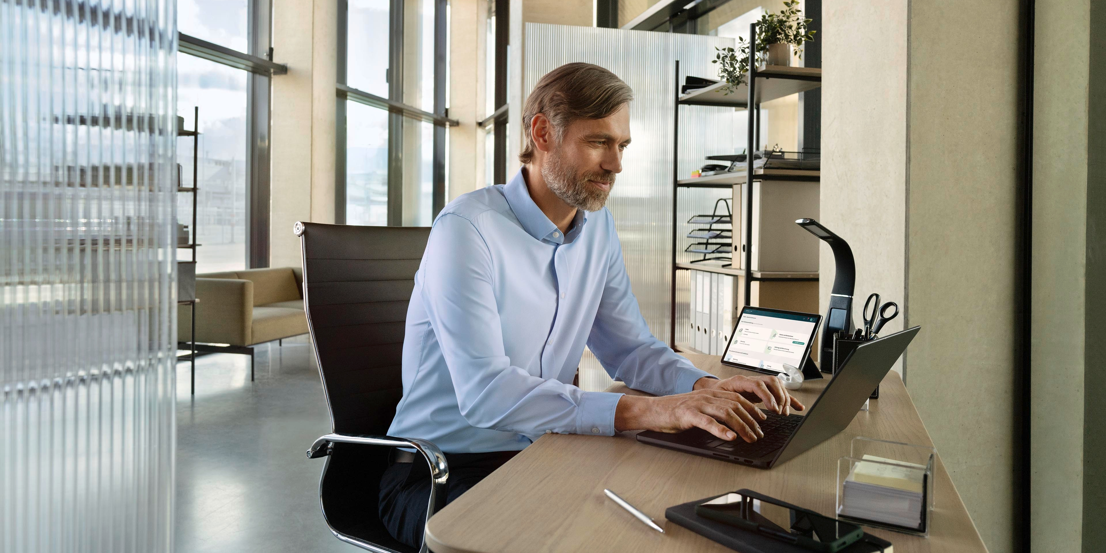 Eine Person sitzt an einem modernen Schreibtisch in einem Büro und arbeitet am Laptop. Im Hintergrund sind große Fenster und Regale mit Büromaterialien zu sehen. Der Raum wirkt hell und gut beleuchtet.