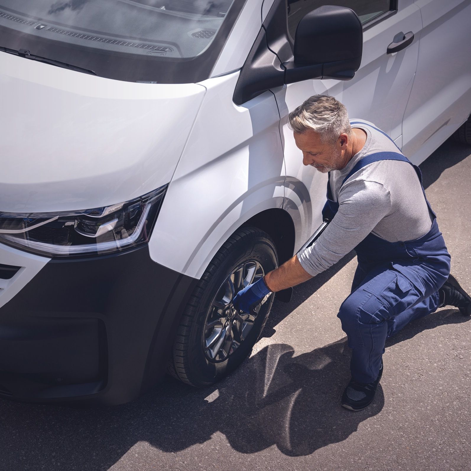 Ein Volkswagen Techniker in blauer Latzhose kniet neben einem weißen VW-Transporter und zieht mit einem Radkreuz die Radmuttern eines Vorderrads an.
