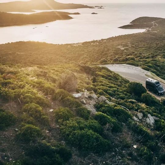 Ein VW Grand California fährt eine kurvige Straße hinauf, umgeben von grüner Vegetation. Im Hintergrund sieht man die Küste mit kleinen Inseln und das Meer. Die Sonne geht über der Szene unter, was zu einer warmen Beleuchtung führt.