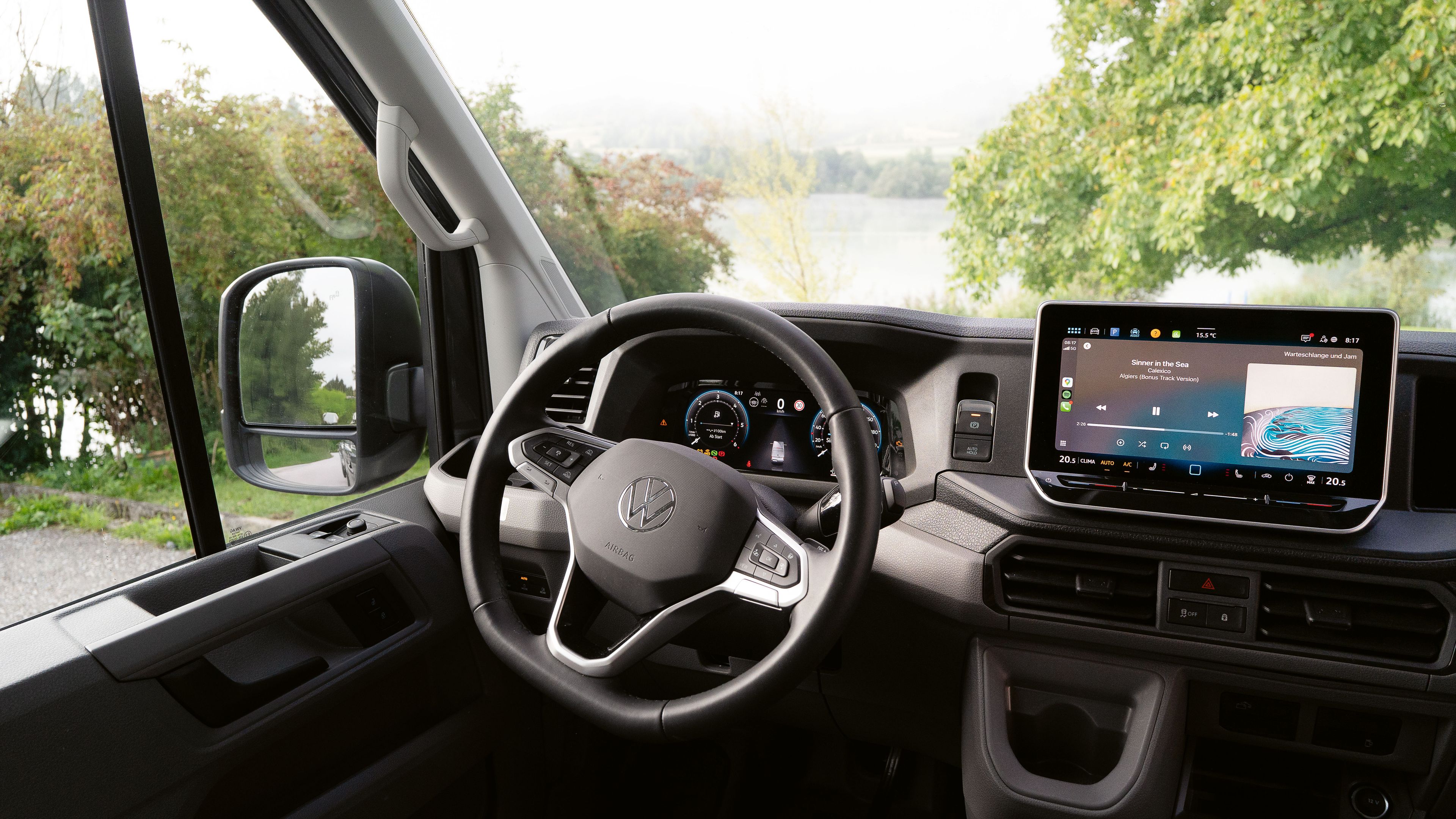 Innenraum eines modernen Autos, das auf einen See blickt. Im Mittelpunkt steht das Lenkrad und ein großes Touchscreen-Display auf dem Armaturenbrett. Durch die Fenster ist eine grüne Landschaft mit Bäumen und Wasser zu sehen.