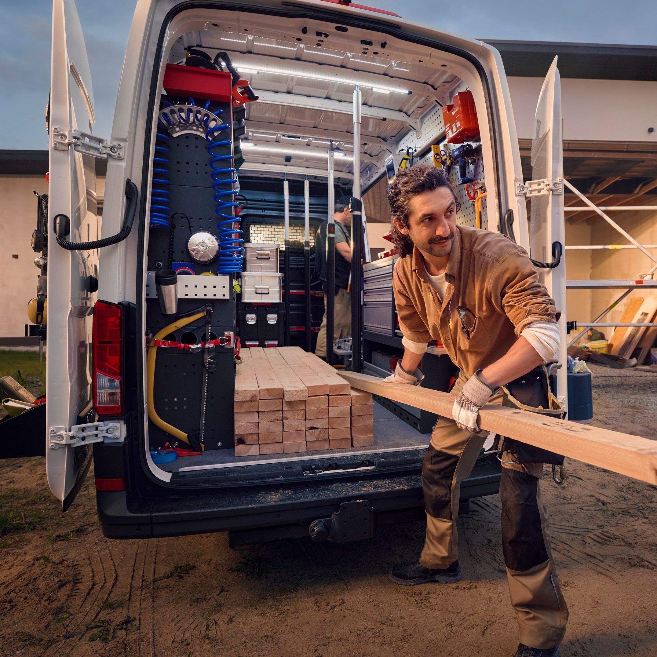 Ein Bauarbeiter lädt Holz aus einem Lieferwagen vor einem Haus, das sich im Bau befindet. Im Hintergrund sind Gerüste und Baumaterialien zu sehen.