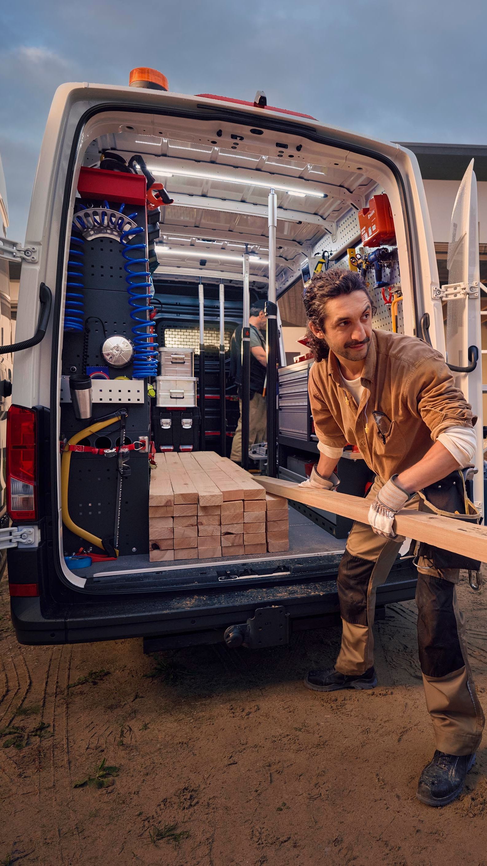 Ein Bauarbeiter lädt Holz aus einem Lieferwagen vor einem Haus, das sich im Bau befindet. Im Hintergrund sind Gerüste und Baumaterialien zu sehen.