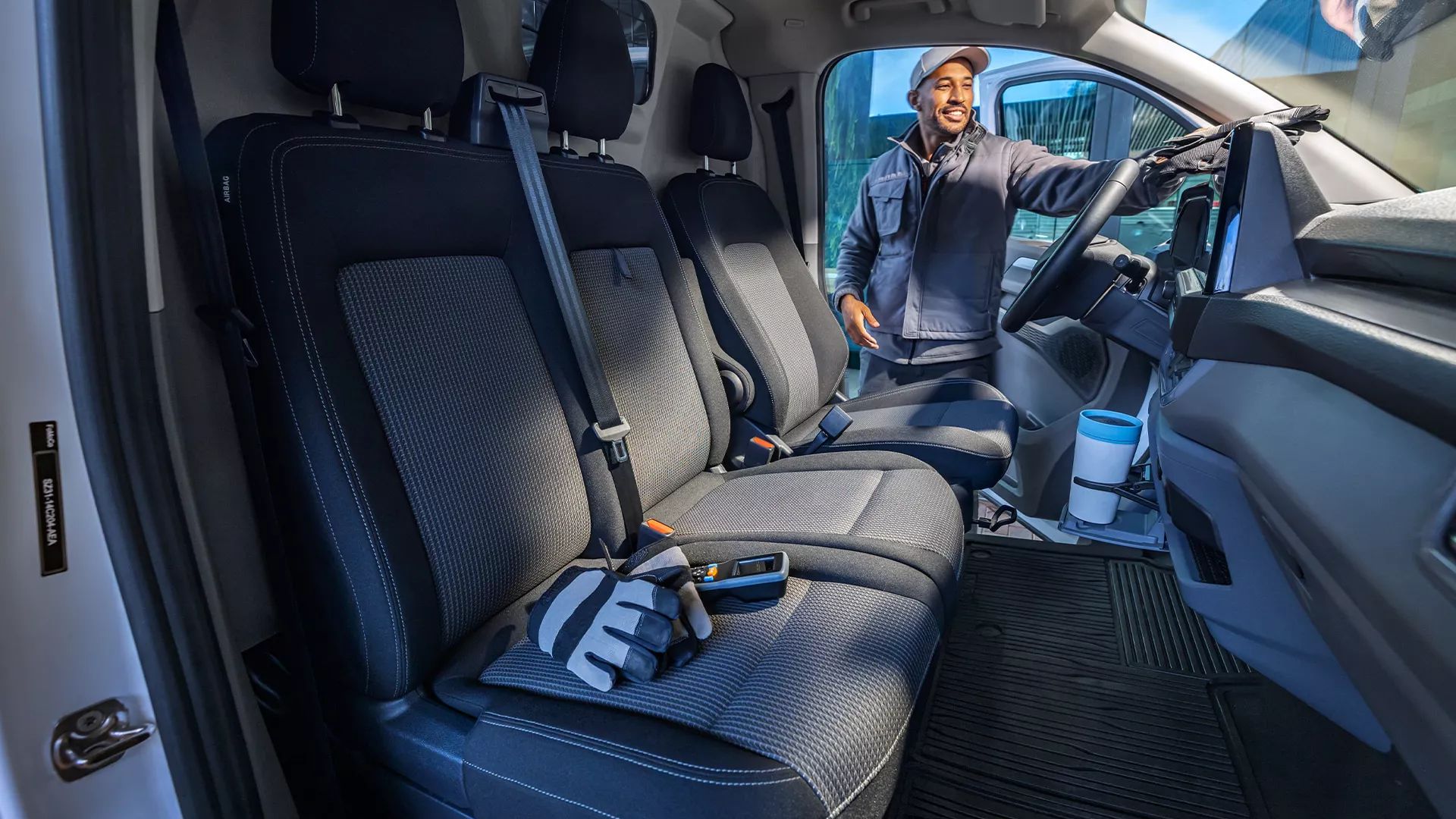 Eine Person steht in geöffneten Tür zur Fahrerkabine des neuen Transporter. Im Vordergrund befindet sich der Innenraum des neuen Transporter mit drei Sitzplätzen und einem Handschuh auf dem Sitz. Die Kabine wirkt sauber und modern.