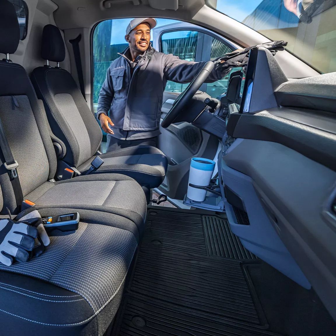 Eine Person steht in geöffneten Tür zur Fahrerkabine des neuen Transporter. Im Vordergrund befindet sich der Innenraum des neuen Transporter mit drei Sitzplätzen und einem Handschuh auf dem Sitz. Die Kabine wirkt sauber und modern.