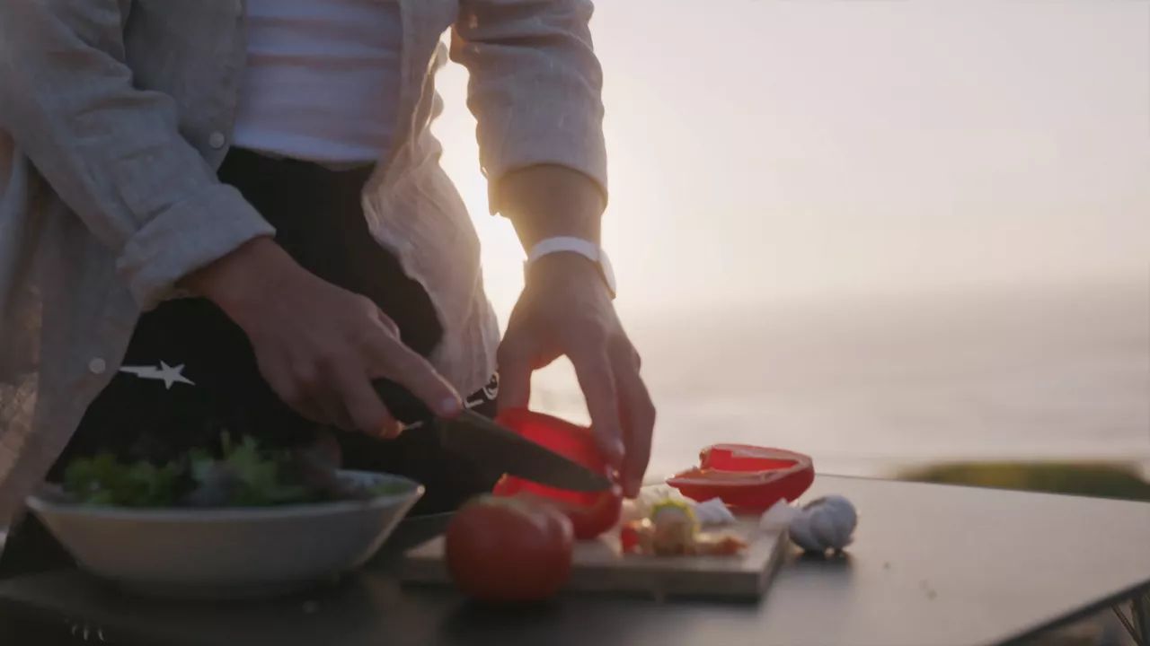 Eine Person schneidet ein rotes Gemüse auf einem Schneidebrett draußen bei Sonnenuntergang. Auf dem Tisch befinden sich weitere Gemüse und eine Schüssel. Im Hintergrund ist eine natürliche Landschaft sichtbar.