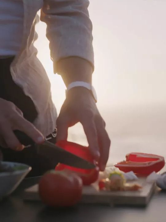 Eine Person schneidet ein rotes Gemüse auf einem Schneidebrett draußen bei Sonnenuntergang. Auf dem Tisch befinden sich weitere Gemüse und eine Schüssel. Im Hintergrund ist eine natürliche Landschaft sichtbar.