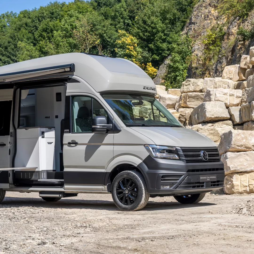 Grand California DUNE steht auf einem steinigen Platz vor einem Hügel mit grünem Bewuchs. Neben dem Wohnmobil sind zwei Campingstühle und ein ausgefahrener Schatten. Das Fahrzeug hat ein erhöhtes Dach und eine Tür, die in den Wohnbereich führt.