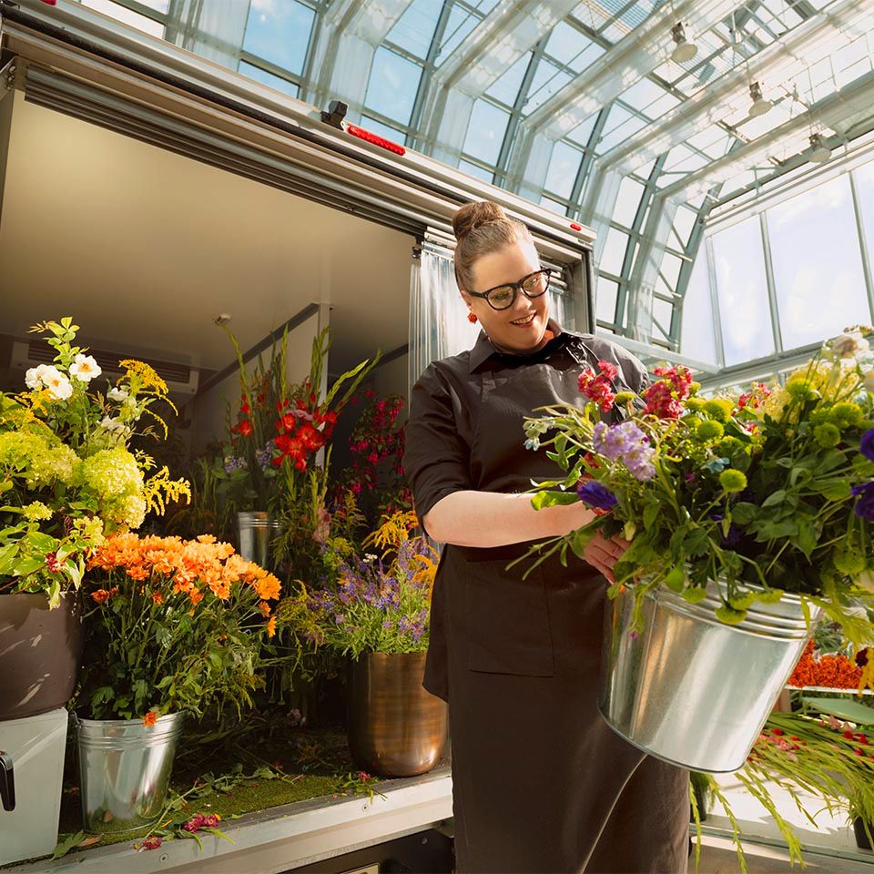 Eine Person steht in einem Blumenladen, umgeben von einer Vielzahl von bunten Blumenarrangements. Sie hält einen silbernen Eimer voller frischer Blumen. Der Raum hat große Fenster, die viel Tageslicht hereinlassen.