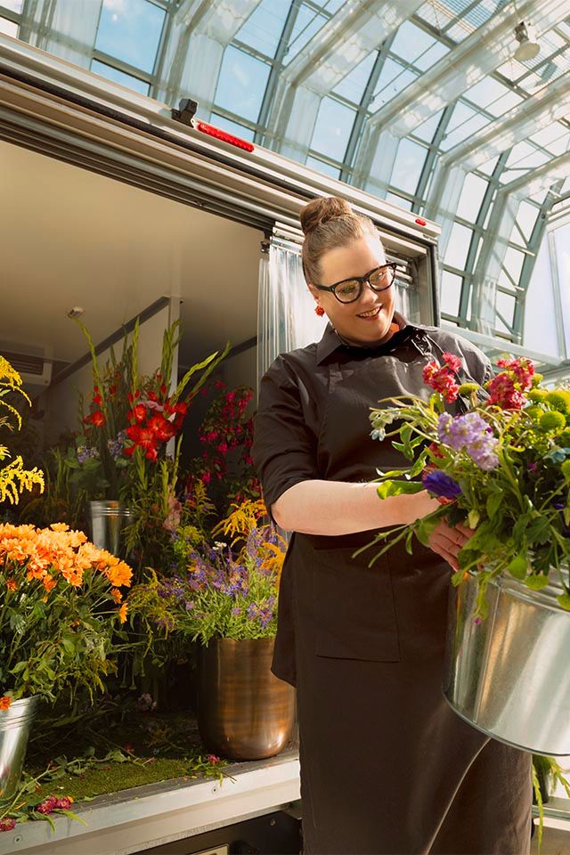Eine Person steht in einem Blumenladen, umgeben von einer Vielzahl von bunten Blumenarrangements. Sie hält einen silbernen Eimer voller frischer Blumen. Der Raum hat große Fenster, die viel Tageslicht hereinlassen.