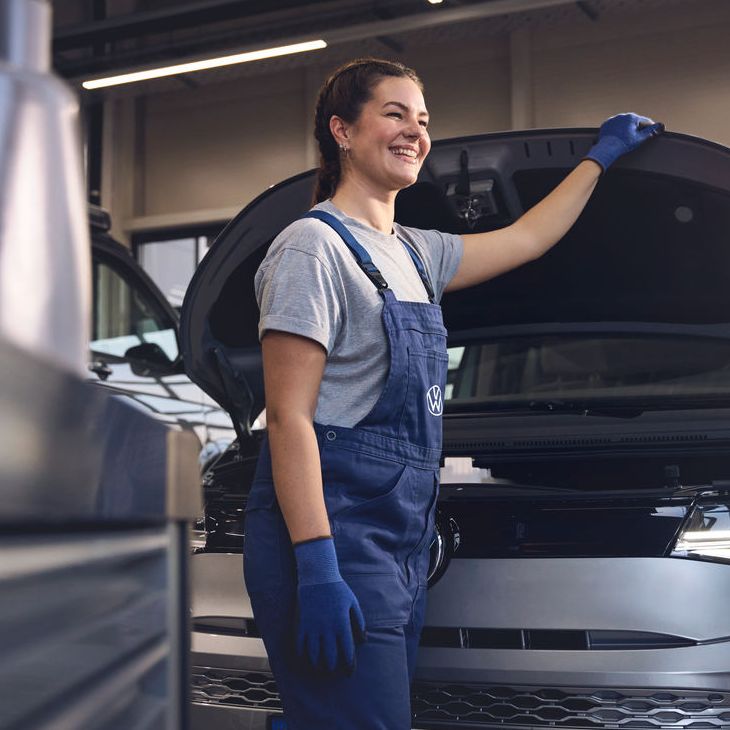 Eine Volkswagen Mechanikerin in blauer Arbeitskleidung steht in einer Volkswagen Werkstatt und inspiziert den Motorraum eines offenen Volkswagen. Sie trägt Handschuhe und stützt sich mit einer Hand auf die Motorhaube.