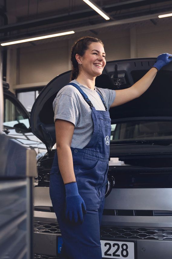 Eine Volkswagen Mechanikerin in blauer Arbeitskleidung steht in einer Volkswagen Werkstatt und inspiziert den Motorraum eines offenen Volkswagen. Sie trägt Handschuhe und stützt sich mit einer Hand auf die Motorhaube.