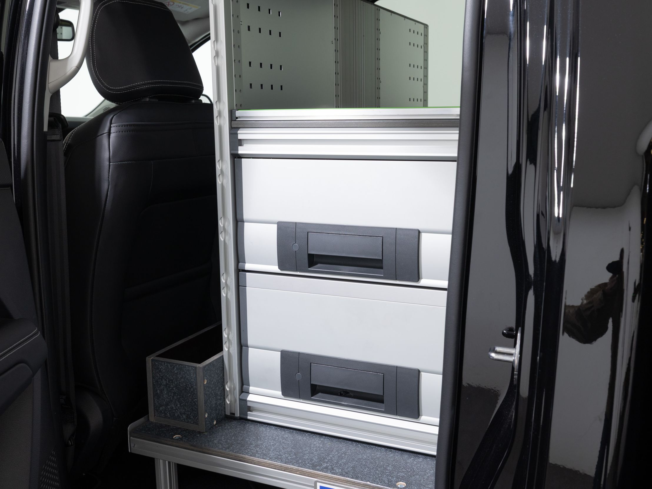 Ein Foto zeigt den Innenraum eines Fahrzeugs mit einer eingebauten Organisations- und Lagerbox. Zwei große Schubladen sind in einem Rahmen verankert, und daneben ist eine kleine Metallbox platziert. Diese Vorrichtung befindet sich hinter dem Fahrersitz.