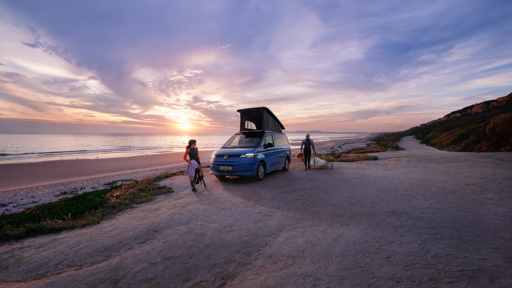 Ein blauer Volkswagen California mit aufgestelltem Dach steht an einem Strand. 