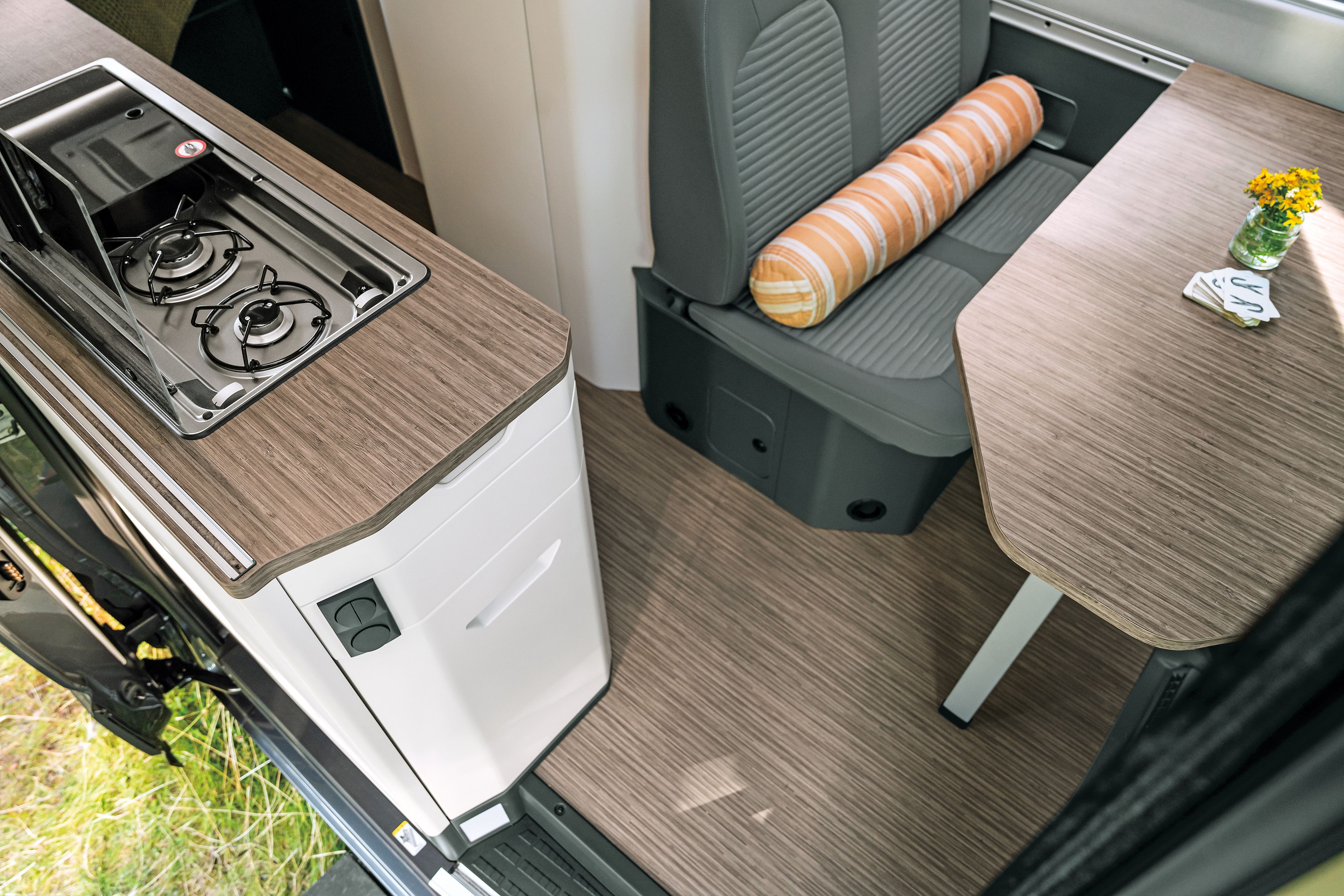 Innenansicht eines Wohnmobils mit einem Gaskocher, einer kleinen Arbeitsfläche und einem Sitzbereich mit einem Tisch. Auf dem Tisch steht eine kleine Vase mit gelben Blumen. Der Sitzbereich ist grau und hat ein gestreiftes Kissen.
