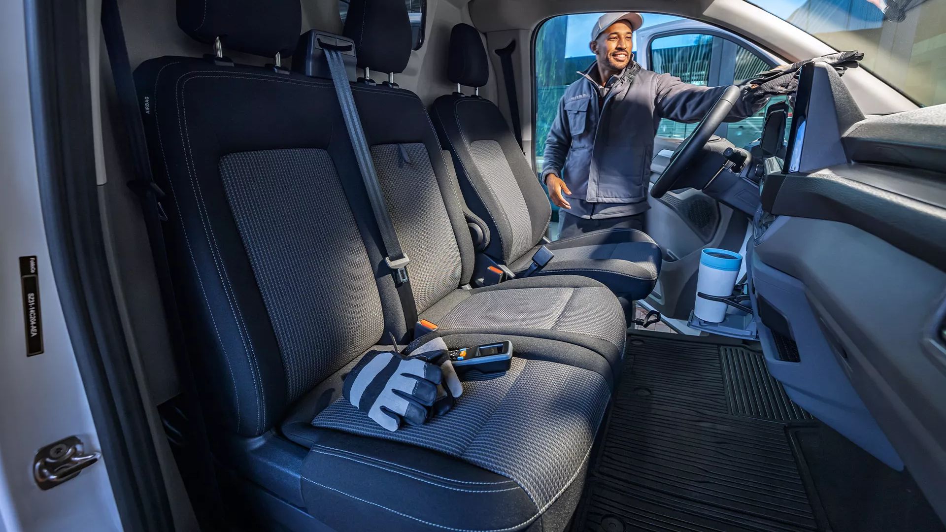 Eine Person steht in geöffneten Tür zur Fahrerkabine des neuen Transporter. Im Vordergrund befindet sich der Innenraum des neuen Transporter mit drei Sitzplätzen und einem Handschuh auf dem Sitz. Die Kabine wirkt sauber und modern.