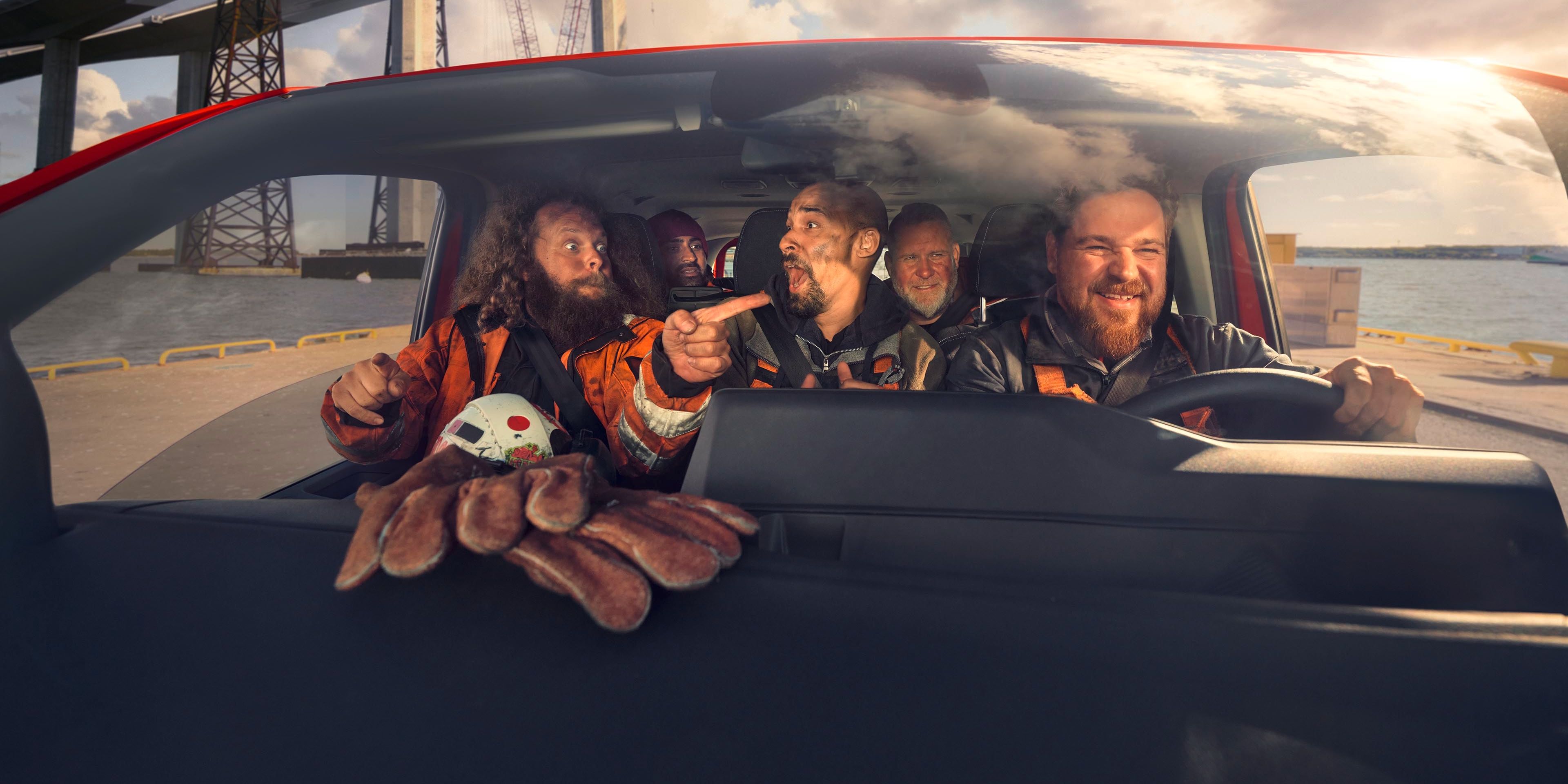 Mehrere Personen in Sicherheitskleidung sitzen in einem Auto und scheinen sich eine Baustelle mit Kranen und Stahlkonstruktionen anzusehen. Eine Person zeigt mit dem Finger in Richtung der Baustelle. Auf der Armaturenbrettablage liegt eine Spielzeugkrabbe.