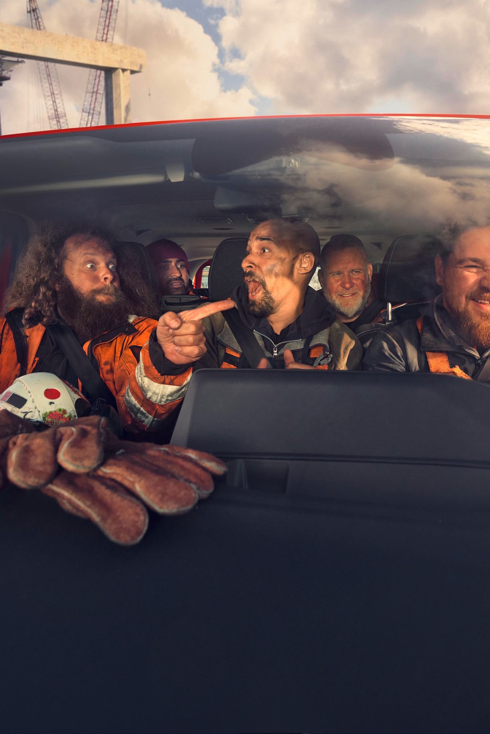 Mehrere Personen in Sicherheitskleidung sitzen in einem Auto und scheinen sich eine Baustelle mit Kranen und Stahlkonstruktionen anzusehen. Eine Person zeigt mit dem Finger in Richtung der Baustelle. Auf der Armaturenbrettablage liegt eine Spielzeugkrabbe.