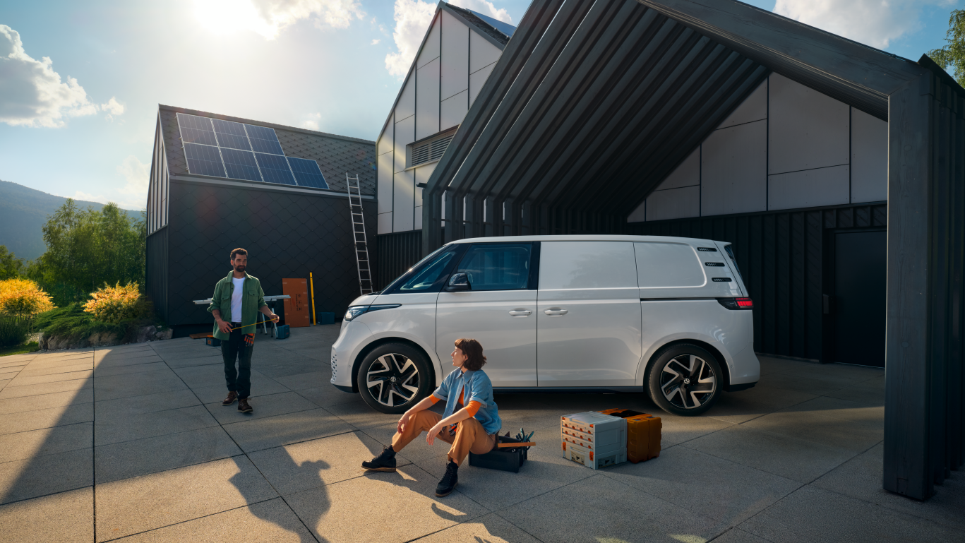 Ein weißer Volkswagen ID. Buzz Cargo vor einer Werkshalle, im Vordergrund sind ein Mann und eine Frau zu sehen