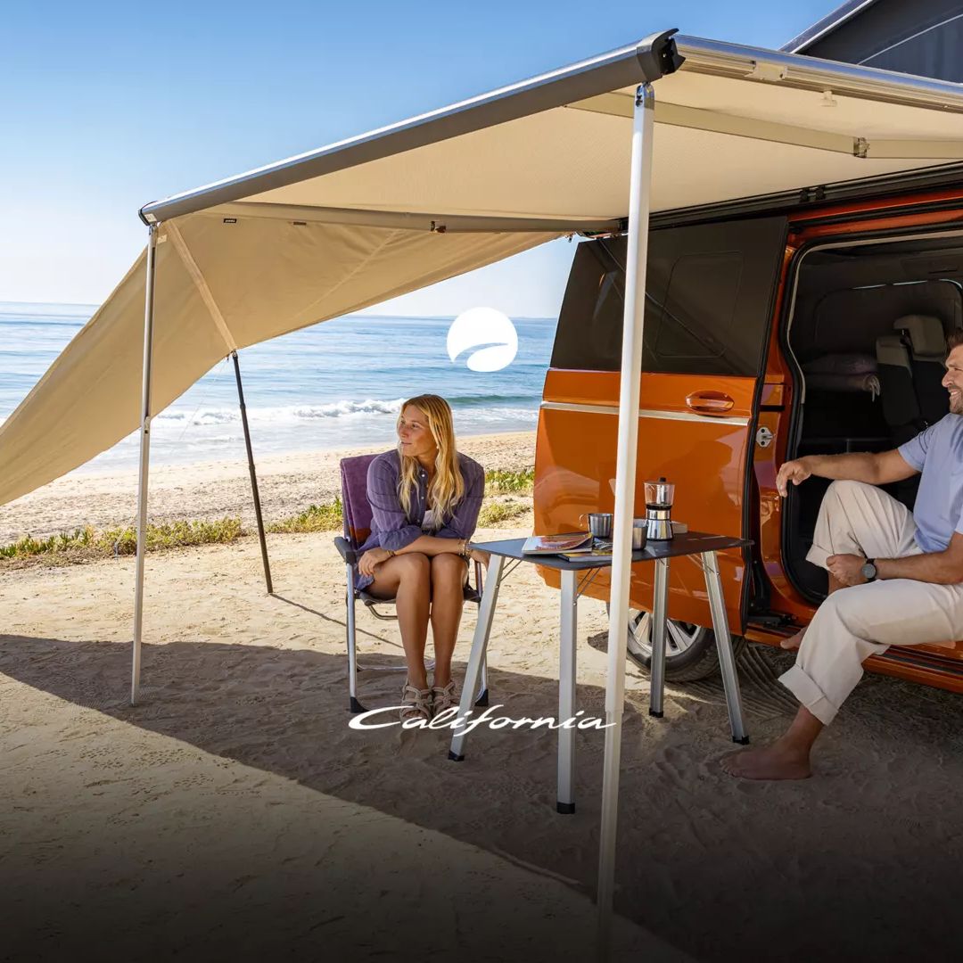 Ein orangefarbener VW California steht am Strand mit ausgefahrenem Sonnendach. Eine Person sitzt im Van, eine andere auf einem Campingstuhl davor. Die Szene zeigt eine entspannte Atmosphäre am Meer.