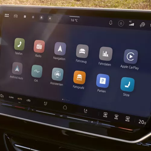 Nahaufnahme eines modernen Infotainment-Touchscreens im Auto mit dunklem Hintergrund und farbigen App-Symbolen. Zu sehen sind Funktionen wie Telefon, Radio, Navigation, Fahrzeug, Fahrdaten, Apple CarPlay, Android Auto und Assistenten. Oben werden Temperatur und Verbindungsstatus angezeigt, unten eine schmale Leiste mit weiteren Bedienelementen.
