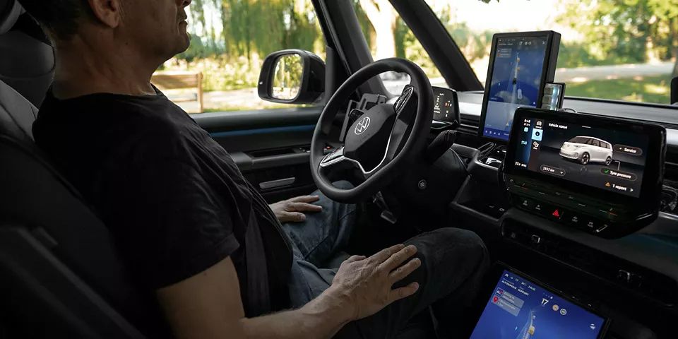 Ein Mann sitzt in einem Auto mit modernen digitalen Armaturen. Das Armaturenbrett zeigt mehrere Bildschirme, die verschiedene Funktionen und Informationen anzeigen. Die Umgebung außerhalb des Autos ist grün und sonnig.