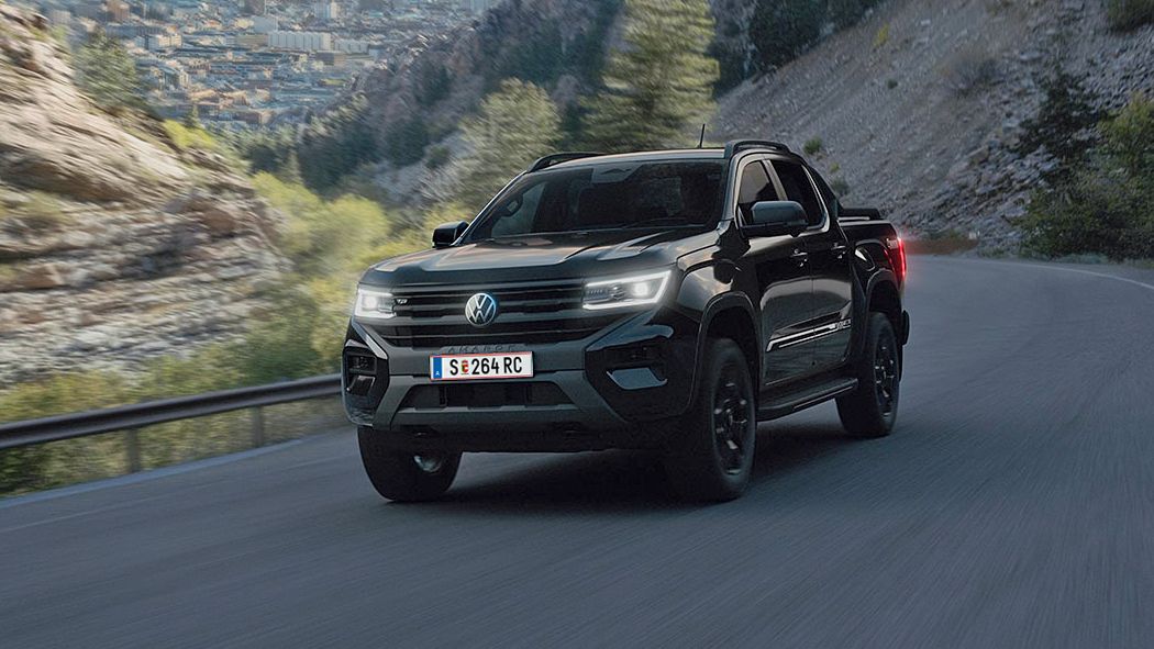 Ein schwarzer Pickup fährt entlang einer kurvenreichen Bergstraße. Im Hintergrund sind steile Felsen und eine Landschaft mit Bäumen und einer Stadt zu sehen. Der Himmel ist teilweise bewölkt.