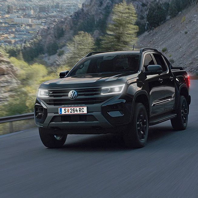 Ein schwarzer Pickup fährt entlang einer kurvenreichen Bergstraße. Im Hintergrund sind steile Felsen und eine Landschaft mit Bäumen und einer Stadt zu sehen. Der Himmel ist teilweise bewölkt.
