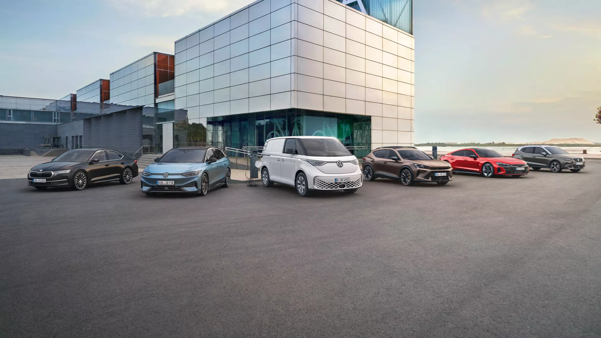 Eine Reihe von Fahrzeugen steht vor einem modernen Gebäude. Es sind verschiedene Autotypen, darunter Limousinen, ein Van und ein Sportwagen. Die Autos sind in unterschiedlichen Farben lackiert.