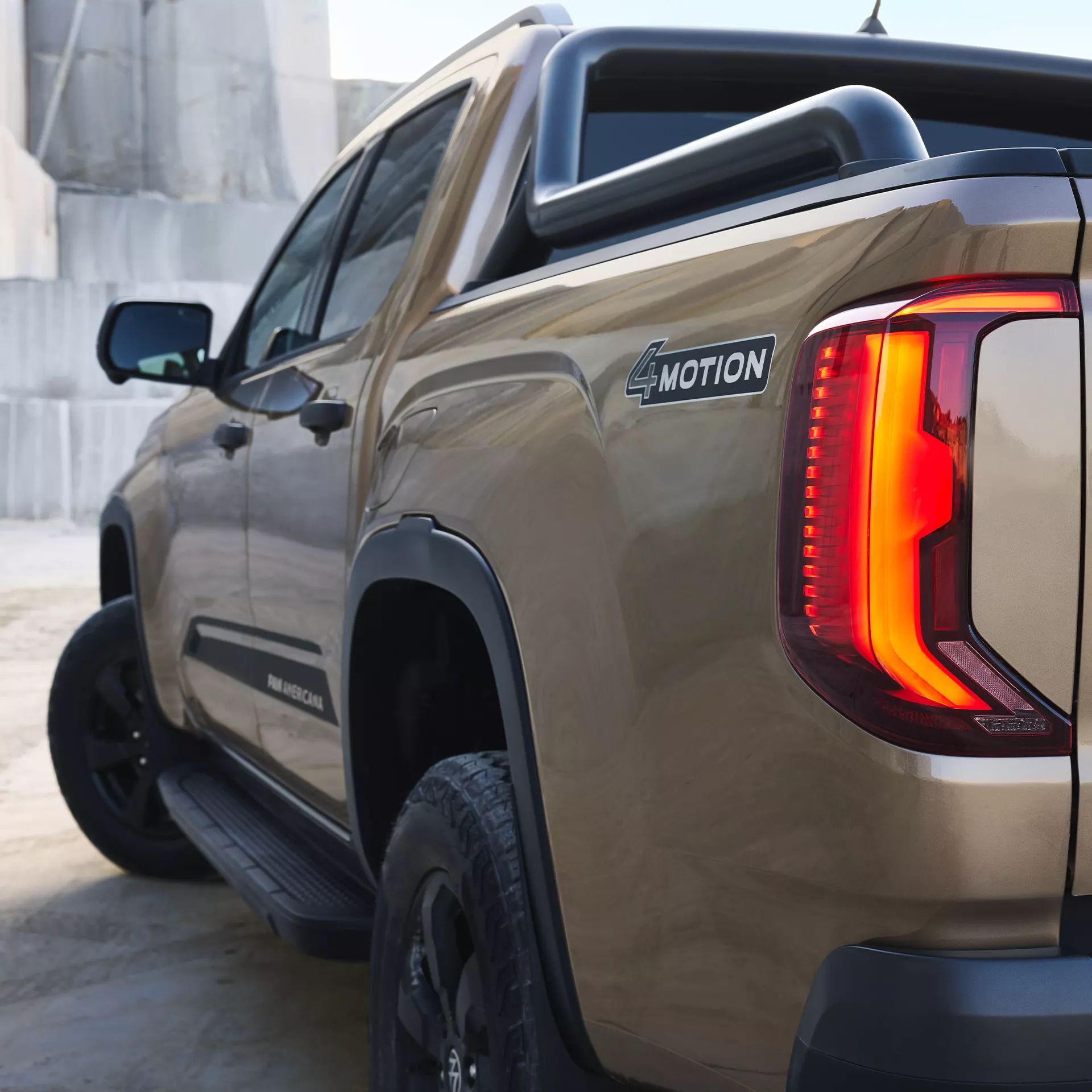 Ein brauner Pickup-Truck ist seitlich und von hinten zu sehen. Auf der Karosserie befinden sich die Schriftzüge "4MOTION" und "Panamericana". Das Rücklicht des Fahrzeugs ist beleuchtet.