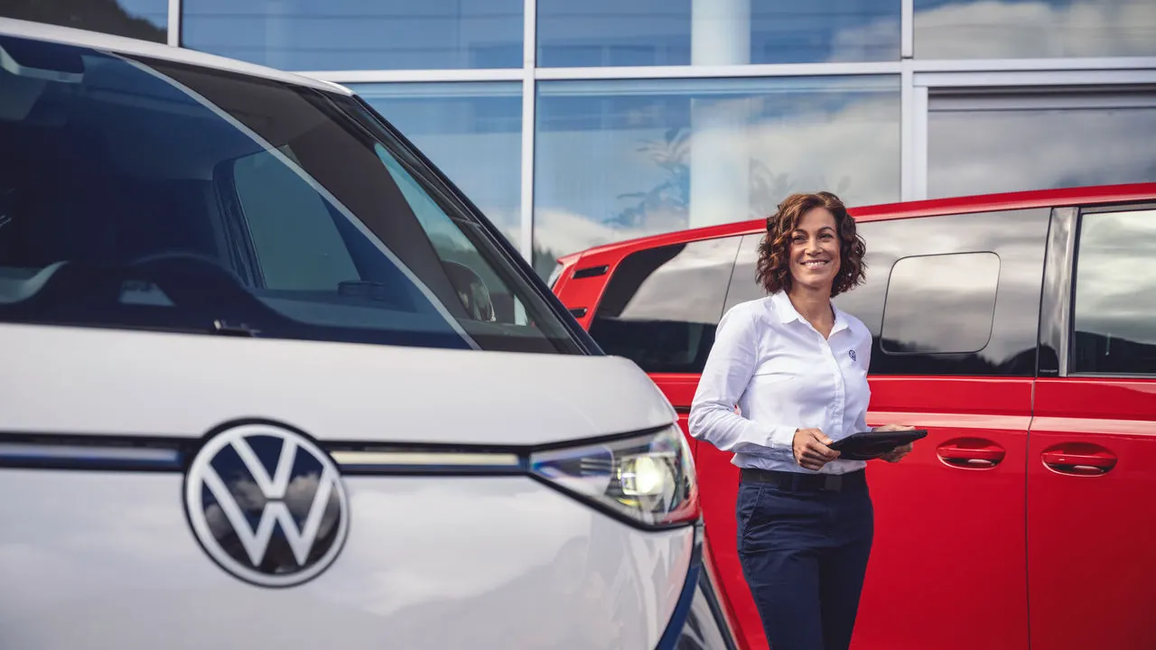 Eine Volkswagen Mitarbeiterin hält ein Tablet in ihren Händen. Links von Ihr steht ein blau weißer und rechts ein roter ID. Buzz.