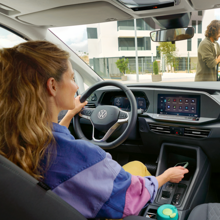Innenraum eines VW Caddy, Frau sitzt am Steuer und legt ihr Handy in die Mittelkonsole, Mann mit Baby geht vor dem Auto