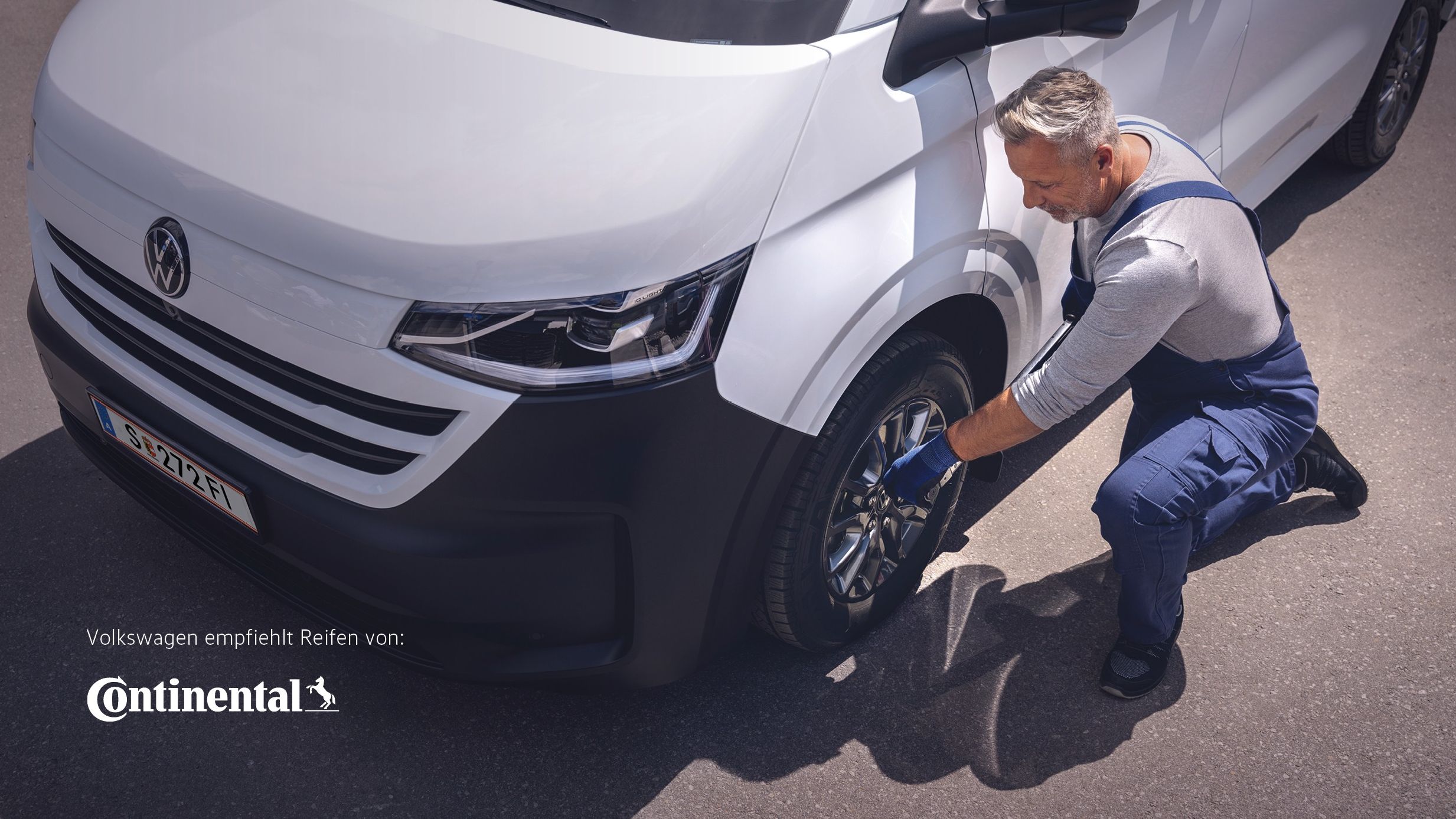 Ein Volkswagen Techniker in blauer Latzhose kniet neben einem weißen VW-Transporter und zieht mit einem Radkreuz die Radmuttern eines Vorderrads an.