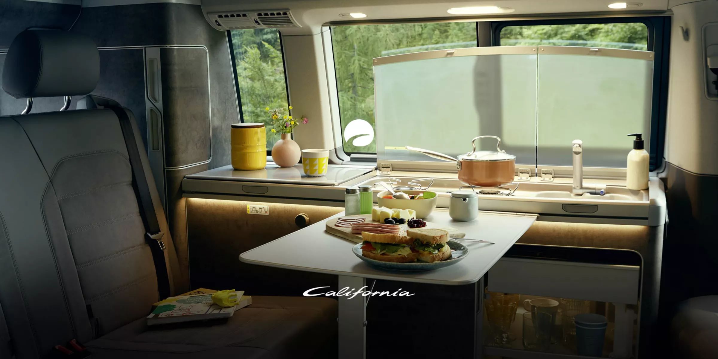 Das Bild zeigt das Innere eines VW California mit einem kleinen Tisch, auf dem Frühstück serviert wird. Im Hintergrund sieht man das Spülbecken sowie sonstige Küchenutensilien. Durch das Fenster ist viel Grün zu erkennen.