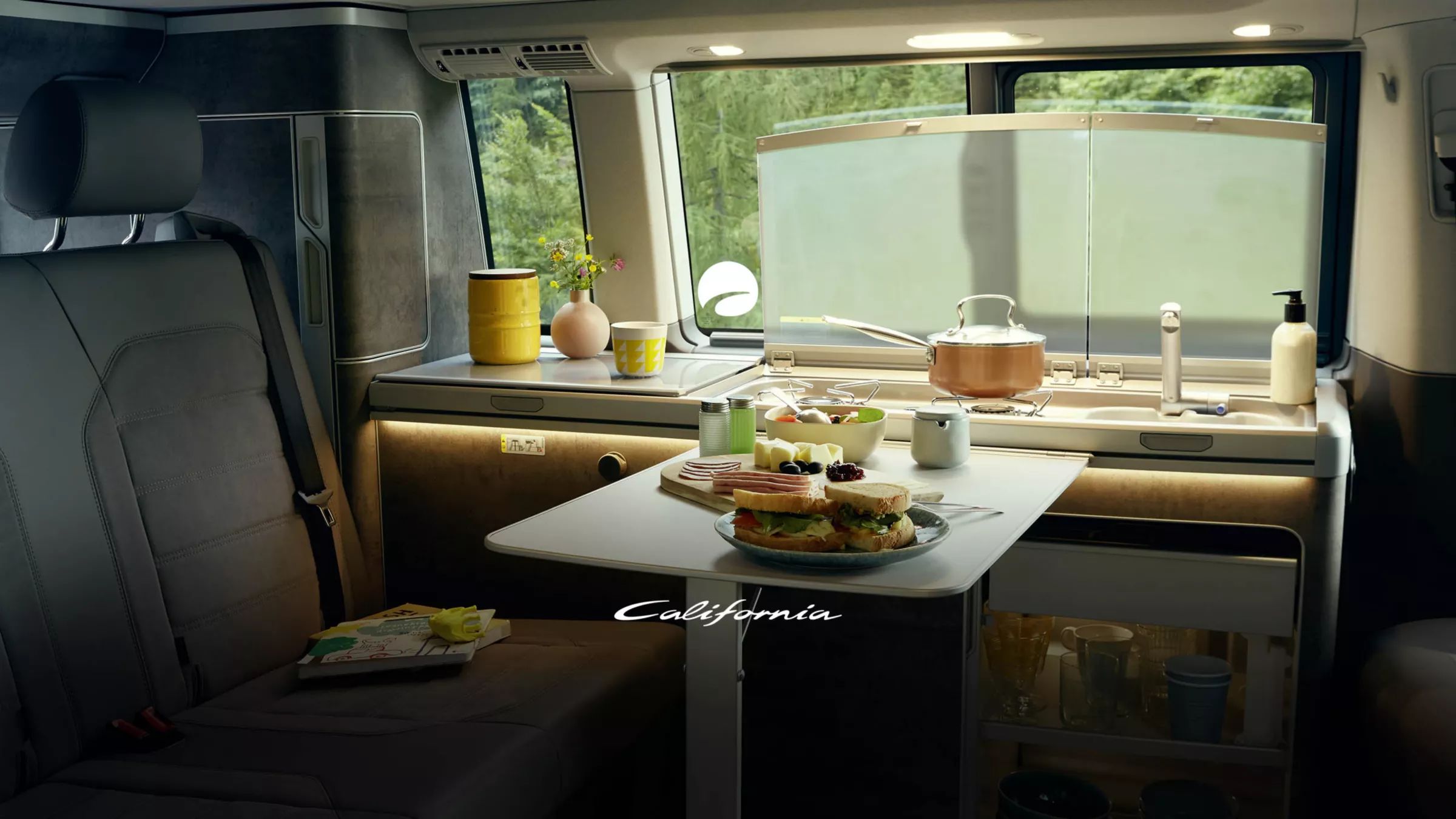 Das Bild zeigt das Innere eines VW California mit einem kleinen Tisch, auf dem Frühstück serviert wird. Im Hintergrund sieht man das Spülbecken sowie sonstige Küchenutensilien. Durch das Fenster ist viel Grün zu erkennen.