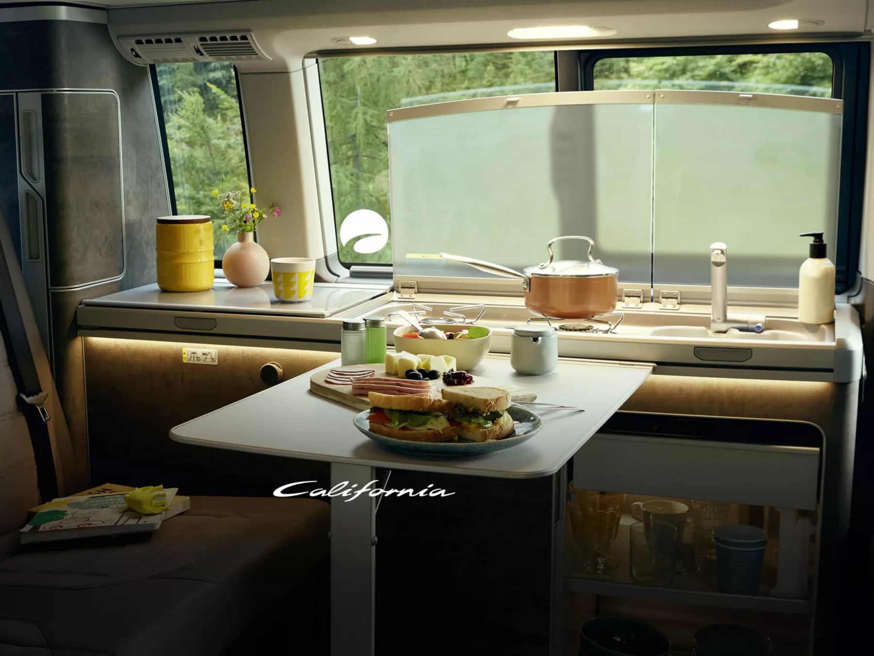 Das Bild zeigt das Innere eines VW California mit einem kleinen Tisch, auf dem Frühstück serviert wird. Im Hintergrund sieht man das Spülbecken sowie sonstige Küchenutensilien. Durch das Fenster ist viel Grün zu erkennen.