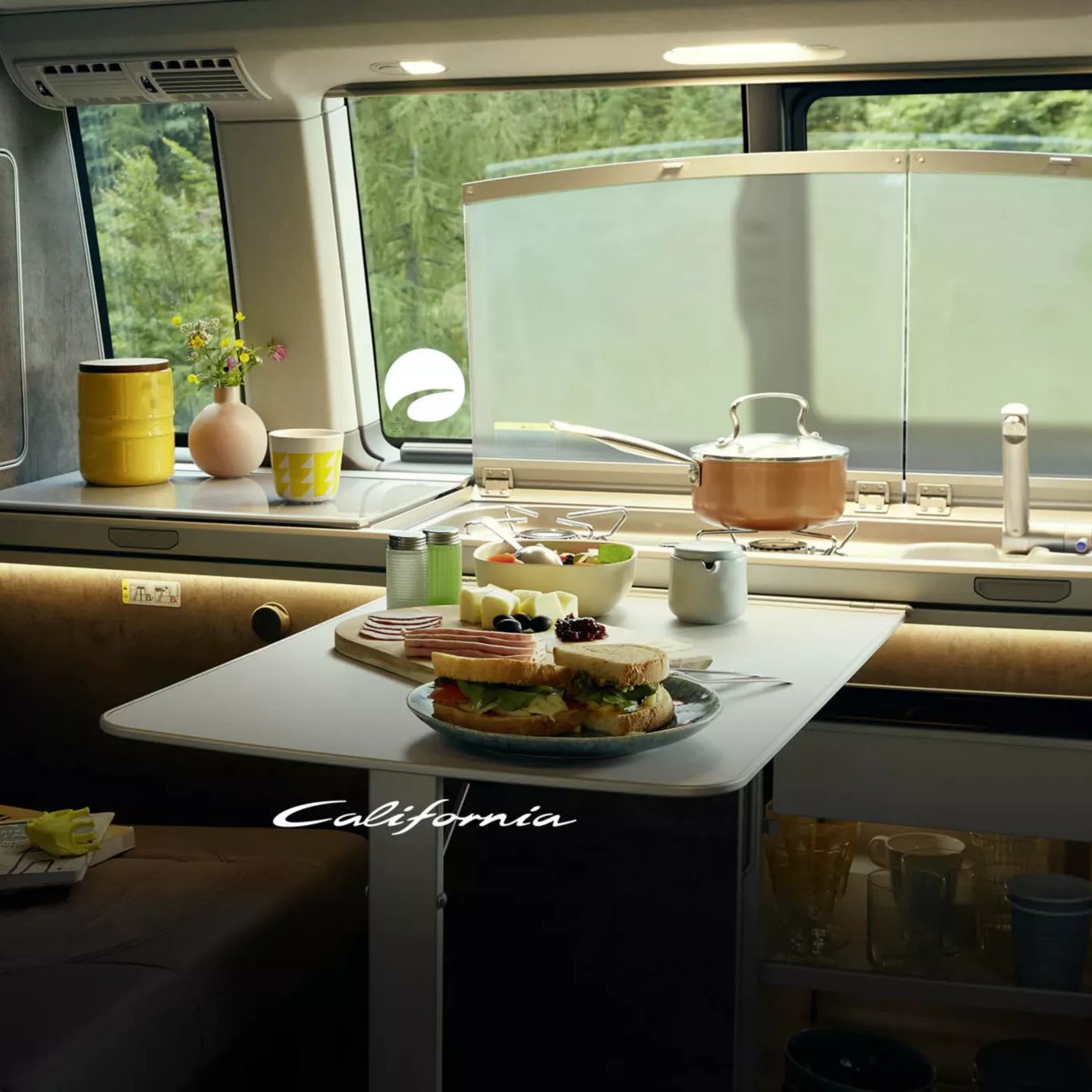 Das Bild zeigt das Innere eines VW California mit einem kleinen Tisch, auf dem Frühstück serviert wird. Im Hintergrund sieht man das Spülbecken sowie sonstige Küchenutensilien. Durch das Fenster ist viel Grün zu erkennen.