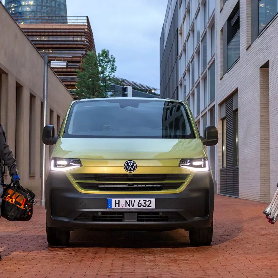Der neue Transporter in grün, steht zwischen modernen Gebäuden auf gepflasterter Straße. Zwei Handwerker stehen links und rechts vom Auto; einer trägt Kabel, der andere eine Leiter. Im Hintergrund ist ein markantes, eiförmiges Gebäude zu sehen.