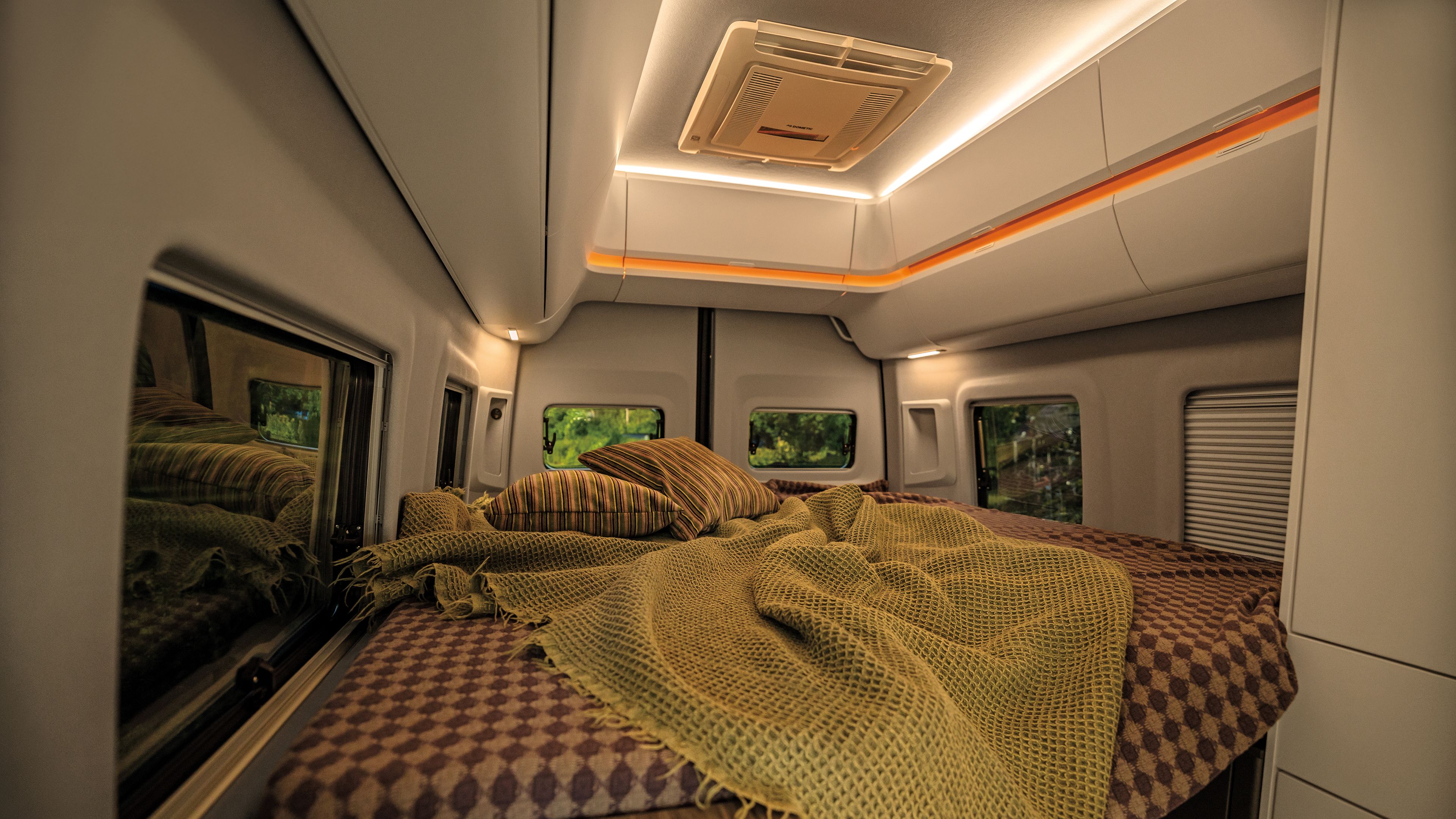 Das Bild zeigt das Interieur eines Wohnmobils mit einem gemütlichen Bett, das mit braun-gemusterten Bettlaken und gelblichen Decken bedeckt ist. An den Seiten sind Fenster zu sehen, durch die grüne Landschaft sichtbar ist. Oben an der Decke befinden sich Beleuchtungselemente.