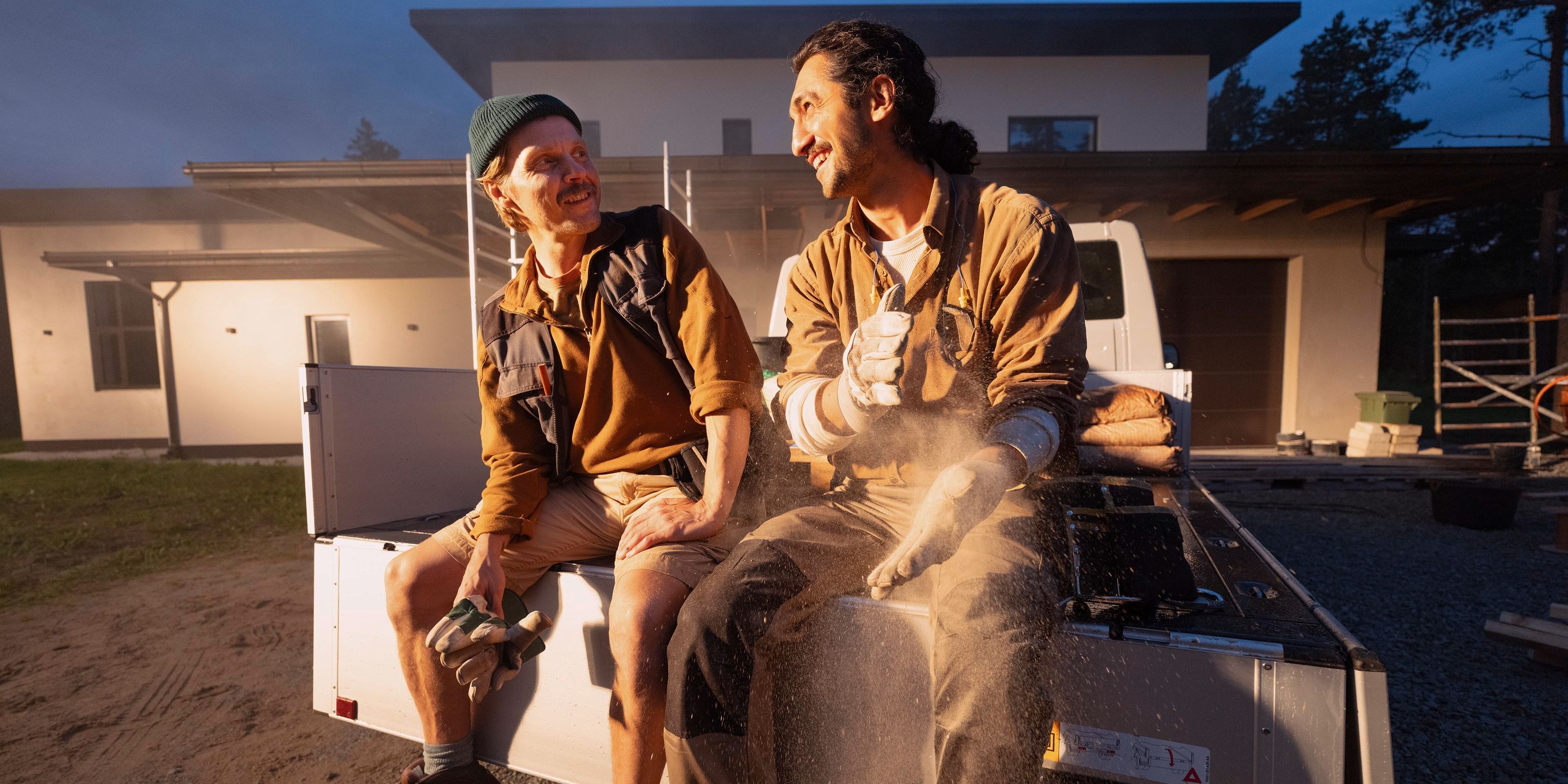 Zwei Personen sitzen auf der Ladefläche eines Pick-up Trucks vor einem modernen Gebäude. Sie tragen Arbeitskleidung und schützen sich vor Staub. Die Umgebung scheint eine Baustelle zu sein.