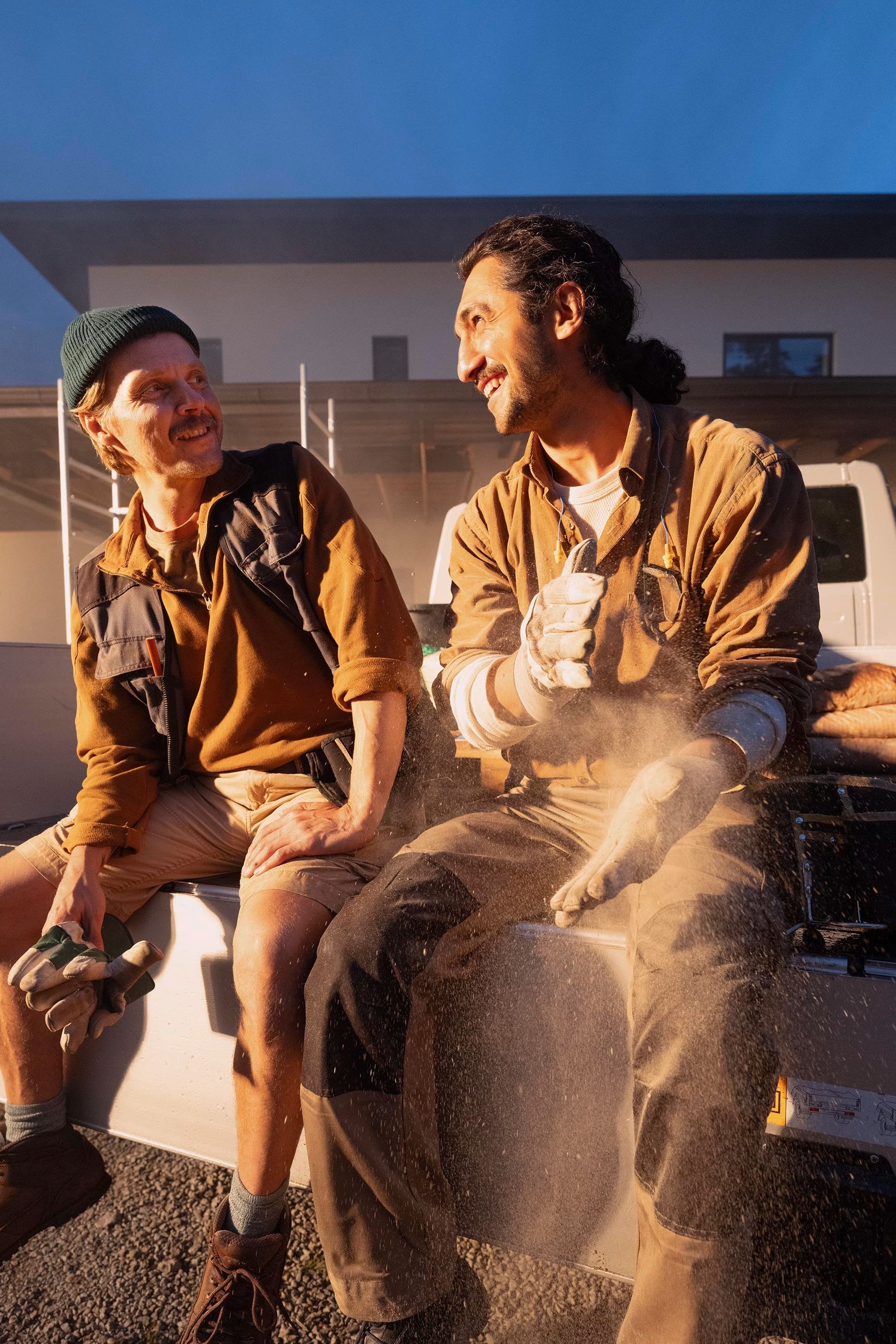 Zwei Personen sitzen auf der Ladefläche eines Pick-up Trucks vor einem modernen Gebäude. Sie tragen Arbeitskleidung und schützen sich vor Staub. Die Umgebung scheint eine Baustelle zu sein.