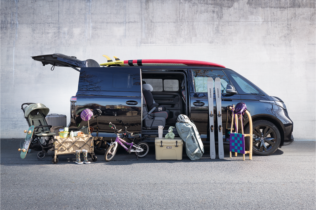 Ein schwarzer VW-Bus steht geparkt vor einer Betonwand, mit geöffneter Seitentür und Kofferraum. Vor dem Bus sind Sport- und Freizeitgegenstände wie ein Kinderfahrrad, ein Skateboard, Skier und ein Schlitten aufgereiht. Auf dem Dach des Busses befinden sich Surfbretter.
