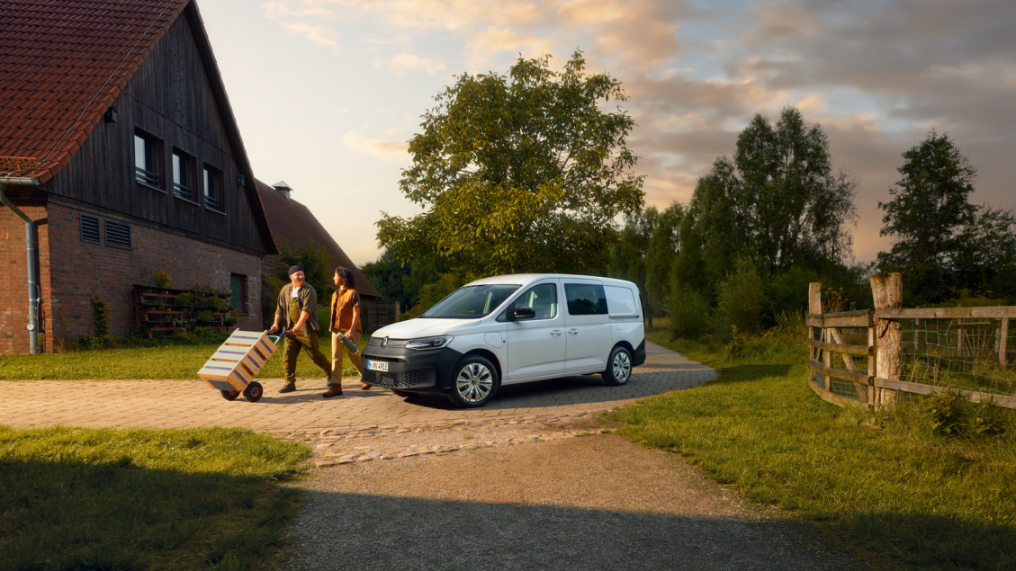 Ein weißer Caddy parkt bei einem Bauernhof. Ein Mann transportiert Kisten mit einer Rodel und eine Frau begleitet ihn.