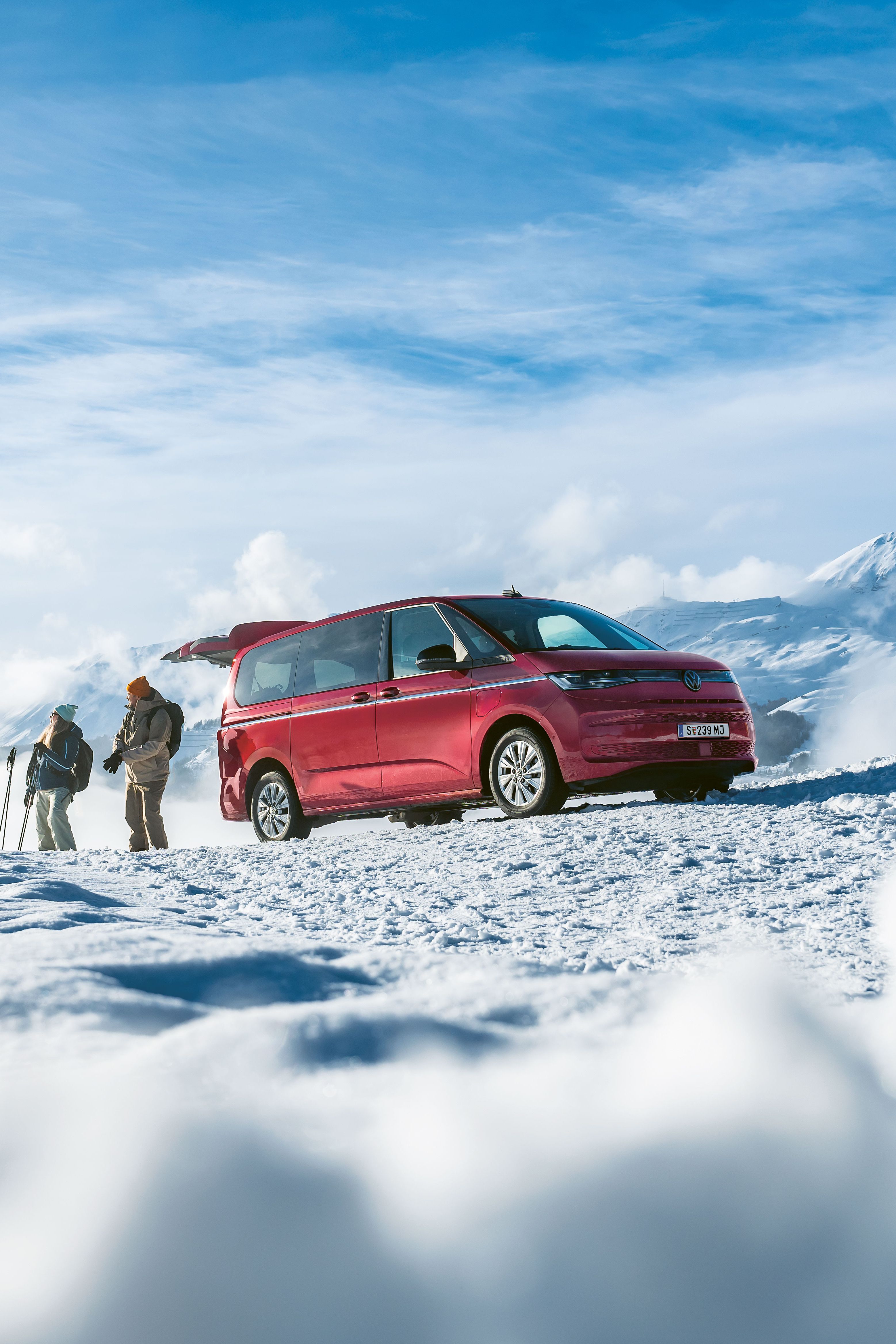 Ein roter Volkswagen Multivan mit geöffneter Heckklappe parkt in den verschneiten Bergen. Ein Mann und eine Frau mit Skiern stehen davor.