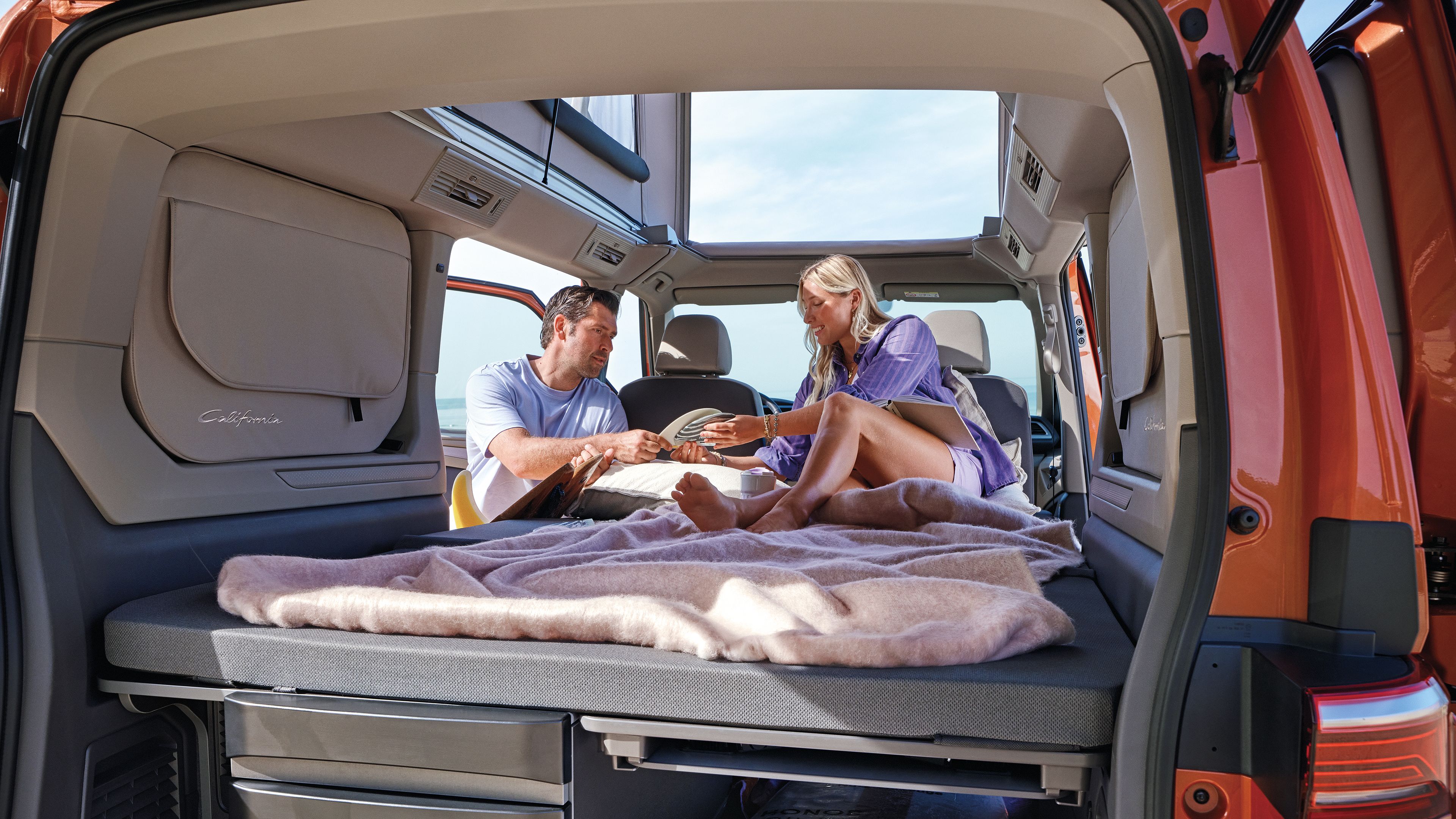 Mann und Frau sitzen auf der Liegefläche eines VW Californias, im Stauraum liegen Surfsachen