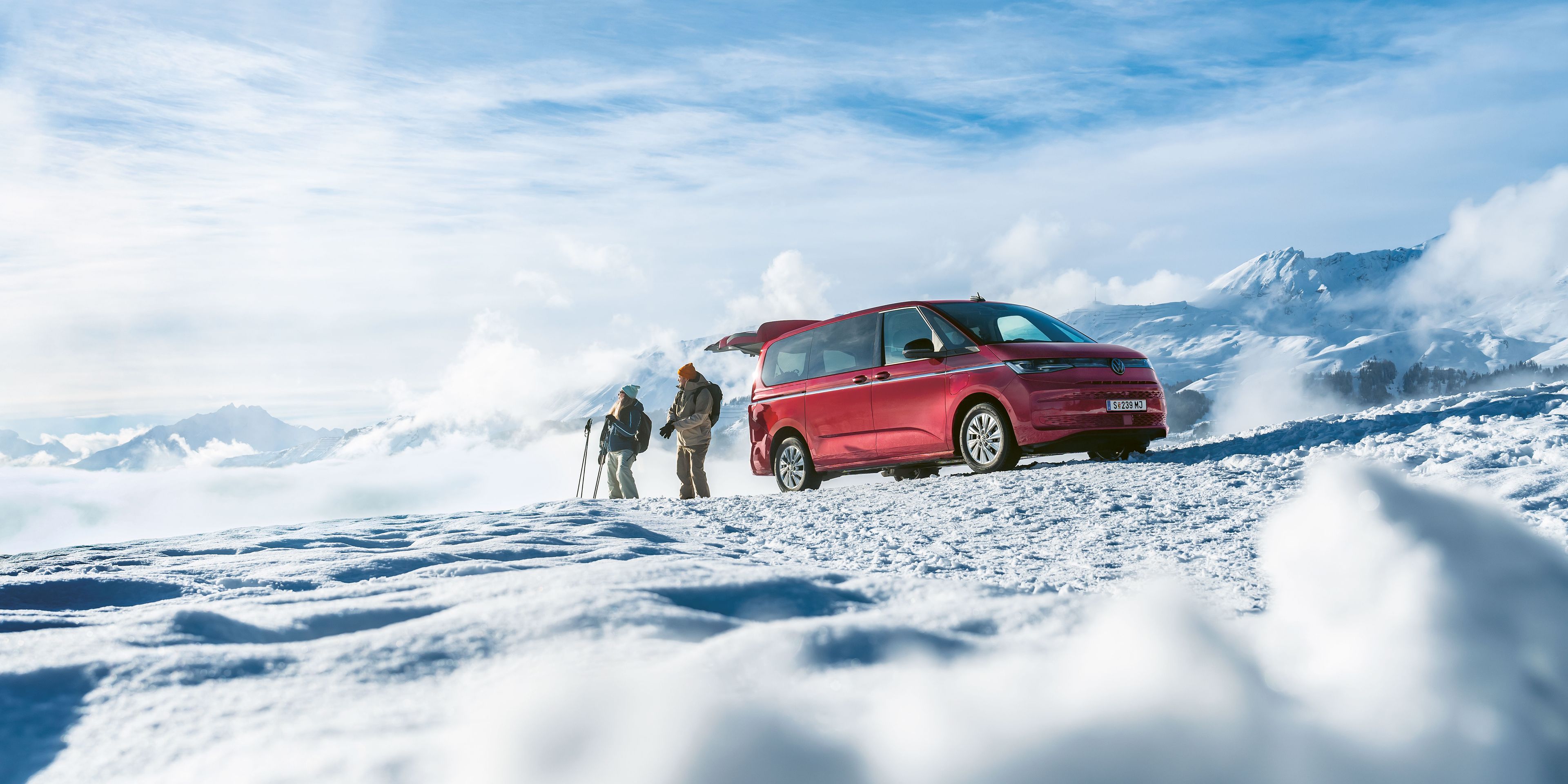 Ein roter Volkswagen Multivan mit geöffneter Heckklappe parkt in den verschneiten Bergen. Ein Mann und eine Frau mit Skiern stehen davor.