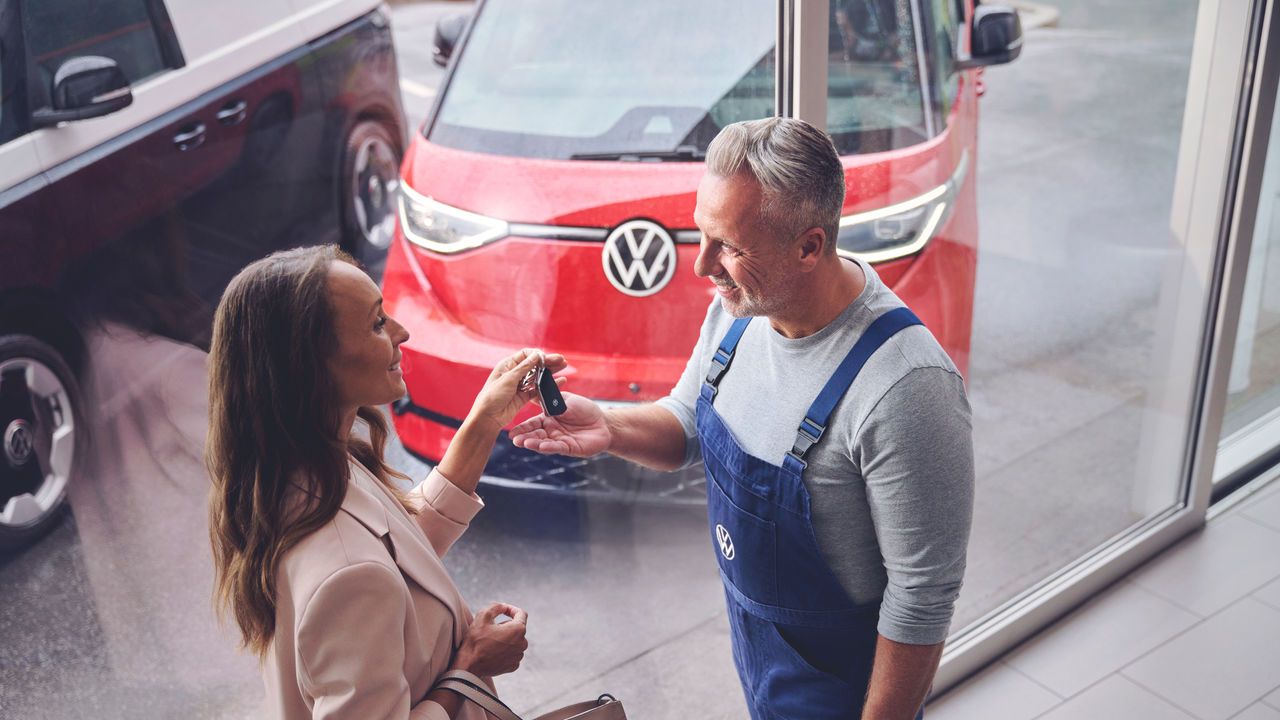 Ein Volkswagen Techniker überreicht einer Kundin ihren Autoschlüssel. Im Hintergrund befindet sich ein roter Volkswagen ID. Buzz.