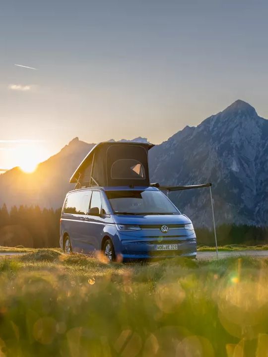 Ein blauer Campingbus steht auf einer grünen Wiese, die Sonne geht hinter einem bewaldeten Berghang unter. Das Dach des Campingbusses ist aufgeklappt. Im Hintergrund sind hohe Berge zu sehen.